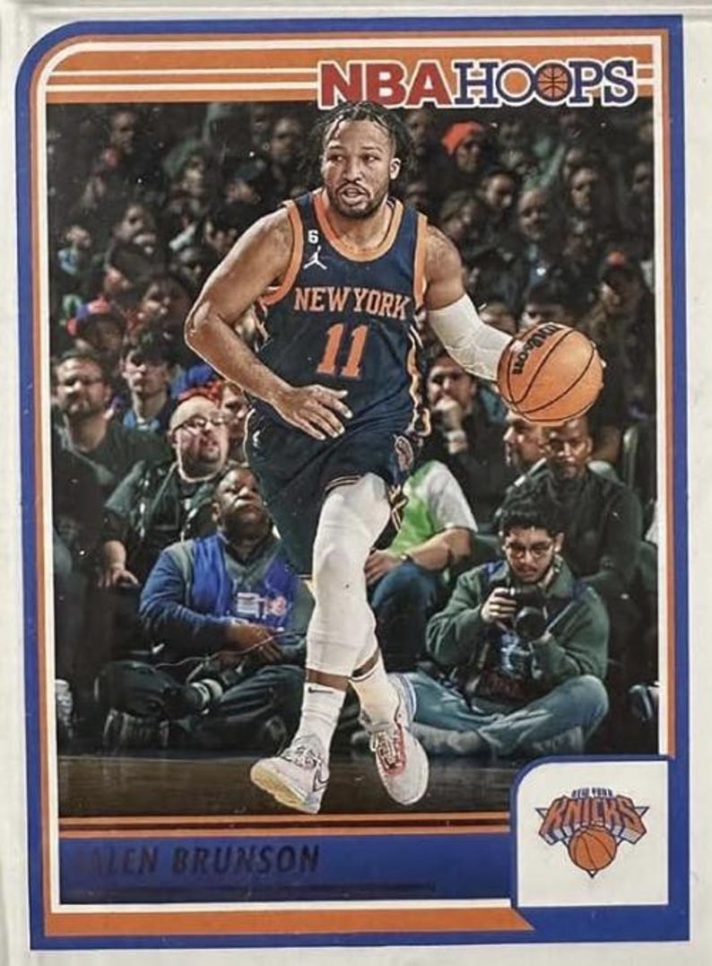 Puede incluir: Una tarjeta de intercambio de baloncesto con un jugador con un jersey azul y naranja de los New York Knicks con el n&uacute;mero 11 en la parte delantera. El jugador est&aacute; botando un bal&oacute;n de baloncesto y lleva zapatillas blancas. La tarjeta tiene un borde azul y naranja con las palabras "NBA Hoops" en la parte superior y "Jalen Brunson" en la parte inferior.