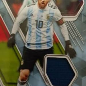 May include: A Panini trading card featuring Lionel Messi in an Argentina national team jersey. The jersey is white and light blue with the number 10. Messi is wearing black shorts and gloves, with a soccer ball at his feet.