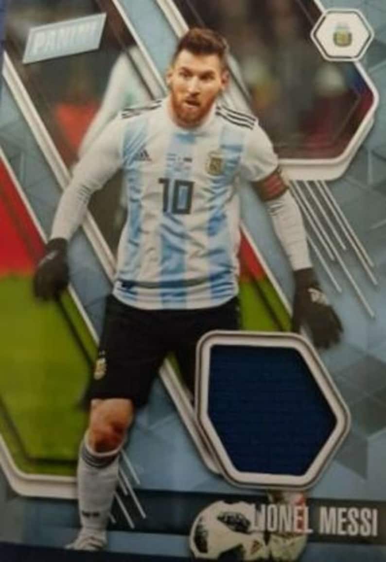 May include: A Panini trading card featuring Lionel Messi in an Argentina national team jersey. The jersey is white and light blue with the number 10. Messi is wearing black shorts and gloves, with a soccer ball at his feet.