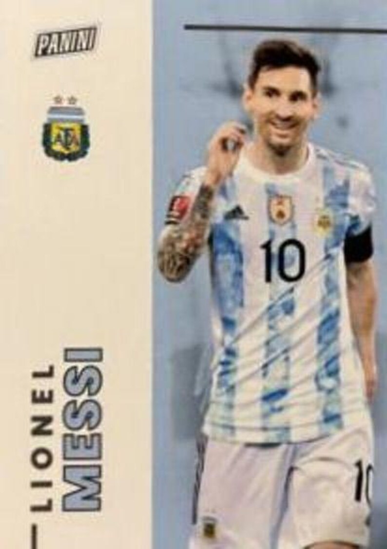 May include: A Panini trading card featuring Lionel Messi in an Argentina national team jersey. The jersey is white with light blue stripes and the number 10. The card also displays the Panini logo and the Argentina Football Association emblem.