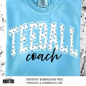 May include: A light blue t-shirt with the words "TEEBALL coach" in white block letters with black polka dots. The shirt is made of a soft material and has a crew neck.
