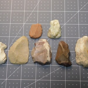 May include: Seven different types of stone tools, including a green stone, a brown stone, a white stone, and a tan stone. The tools are arranged in a semi-circle on a gray surface.