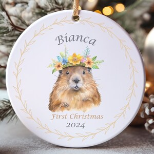 May include: A white ceramic Christmas ornament with a brown and white groundhog wearing a flower crown. The ornament has a gold wreath border and the text "Bianca First Christmas 2024".