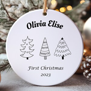 May include: White ceramic Christmas ornament with the names "Olivia Elise" and the text "First Christmas 2023". The ornament features three stylized Christmas trees.
