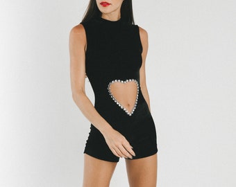 May include: A black velvet jumpsuit with a heart-shaped cutout adorned with silver rhinestones. The jumpsuit has a mock neck and sleeveless design. The model is wearing silver platform sandals.