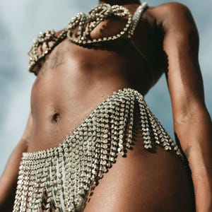 Puede incluir: Un brillante conjunto de bikini plateado. La parte superior es un diseño tipo sujetador, y la parte inferior es una falda con flecos de cristal colgantes. El conjunto está hecho de pedrería brillante. El fondo es un cielo azul claro.