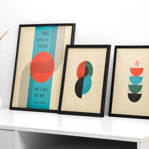 May include: Three framed abstract art prints with geometric shapes and text. The first print has a red circle with black lines and the text "What stands in the way becomes the way". The second print has a green and blue circle with a black circle inside. The third print has a stack of colorful semi-circles.