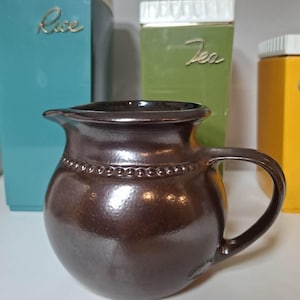 May include: A dark brown ceramic pitcher with a rounded body, a curved handle, and a decorative beaded band around the neck. Behind it are three rectangular kitchen canisters in teal, green, and yellow, labelled Rice and Tea in gold script.