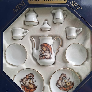 May include: A miniature porcelain tea set with gold trim, comprising a teapot, creamer, sugar bowl, cups, saucers, and a lid. The teapot and saucers display a detailed illustration of a child. The set is presented in a blue box with the text "M.J. Hummel MINI SET".