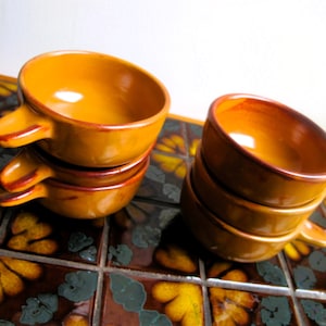 May include: A set of four orange ceramic bowls with handles. The bowls are stacked on top of each other. The bowls are on a tiled surface with a floral pattern.
