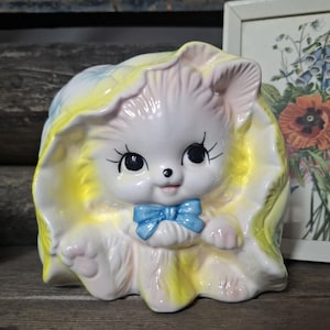 May include: A vintage ceramic cat figurine, white with a blue bow tie, set within a yellow and light blue ruffled design. The figurine has large eyes and a glossy finish. Likely a decorative item, it's reminiscent of mid-century design.
