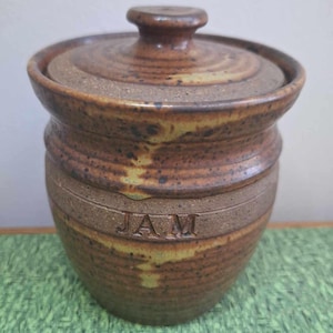 May include: A brown and tan ceramic jar with a lid. The jar has a speckled glaze and the word "JAM" is inscribed on the front.