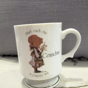 May include: White ceramic mug with a green trim. The mug features a cartoon girl with a red hat and a floral bouquet. The text on the mug reads "Start each day Grandma in a happy way."