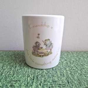 May include: White ceramic tumbler with a charming illustration of a mouse and frog. The tumbler has the text "Friendship is that little something extra..." in a script font. The mouse and frog are dressed in clothes and are around a table.
