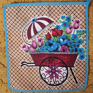 May include: A square pot holder with a brown and white checked pattern, a blue border, and a hanging loop. The central design shows a red wheelbarrow overflowing with blue, red, and purple flowers, and a brown and white umbrella.