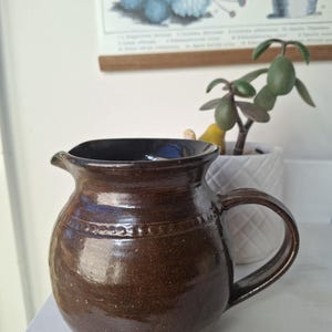 May include: A brown ceramic jug with a handle and spout. The jug has a rounded body and a dark interior. A decorative band of small circles encircles the jug. The jug is likely used for serving beverages.
