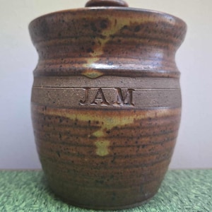 May include: A brown and tan ceramic jar with a lid. The jar has a brown glaze with a speckled pattern and the word "JAM" is inscribed on the front.