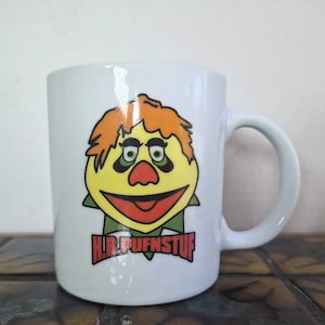 May include: White ceramic mug with a cartoon character design. The character has orange hair, a yellow face, and a red nose and mouth. The mug features the text "H.R. Pufnstuf" in red letters and a green collar.