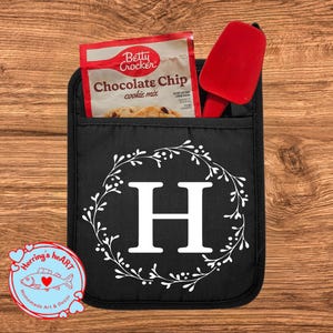 May include: Black oven mitt with a white floral wreath design and the letter "H". A red spatula and a Betty Crocker Chocolate Chip cookie mix packet are in the pocket. A "Herring's heart" sticker is in the lower left corner.
