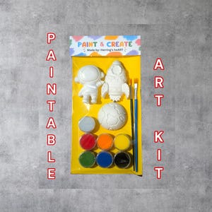 May include: A Paint & Create art kit in a yellow plastic package featuring a white astronaut, rocket, and planet figure. The kit includes two paintbrushes and eight small circular containers of paint in red, orange, yellow, green, blue, and black.