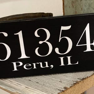 May include: Black wooden sign with white lettering displaying the numbers "61354" and the text "Peru, IL". The sign is resting on a distressed wooden shelf.
