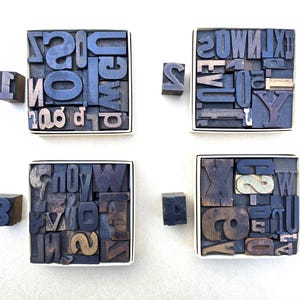 May include: Four square boxes filled with dark wooden letterpress blocks featuring various letters and symbols. Each box is accompanied by a single wooden block marked with the numbers 1, 2, 3, or 4, placed to the left of each respective container.