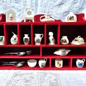 May include: A red wooden display shelf filled with various small, white ceramic figurines and decorative items. The shelf has multiple compartments, and the items include miniature vases, a dog figurine, and spoons.