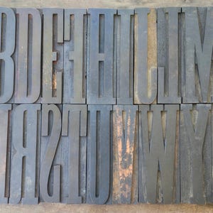 May include: A collection of dark, weathered wooden letterpress blocks arranged in two rows. The top row features the letters A, B, D, E, F, H, I, J, K, L, M, N, and the bottom row displays O, P, Q, R, S, T, U, V, W, Y, and a punctuation mark.