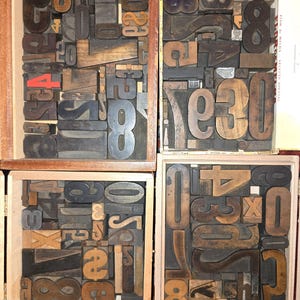 May include: Four wooden boxes filled with an assortment of vintage letterpress printing blocks. The collection includes various numbers, letters, and symbols in different sizes and wood tones, with one block featuring a bright red number four.