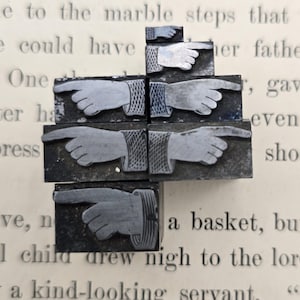 May include: A collection of six metal letterpress printing blocks, each featuring a raised relief of a hand with an extended index finger pointing to the right. The blocks vary in size and detail, showcasing different cuff designs and hand styles.