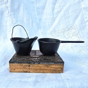 May include: Two small black cast iron pots, one with a handle and the other with a wire loop handle, are sitting on a wooden box with a carved design.