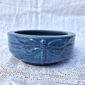 May include: A blue ceramic planter with a raised butterfly design. The planter has a speckled glaze and a rounded shape.