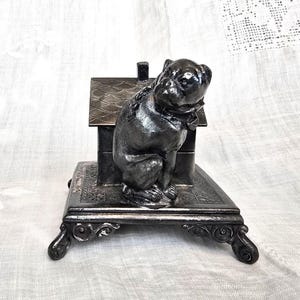 May include: A silver-toned metal figurine of a pug dog sitting in a house-shaped structure. The figurine is on a base with ornate details.