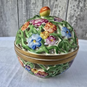 May include: A white ceramic trinket box with a lid. The box is decorated with a floral design of green leaves and colorful flowers. The lid has a small knob on top.