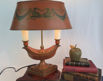 Op de afbeelding: Een vintage tafellamp met twee lampen en een bruine, geverfde metalen kap met een gouden en groen zwanenontwerp. De lampvoet is een gestileerde, goud geverfde metalen olielamp met een vierkante voet.