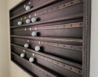 May include: A dark stained wooden display case with multiple horizontal shelves, each holding golf balls. The case has a rectangular frame and is mounted on a wall. The golf balls are white with various markings.