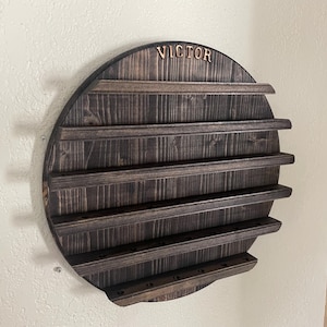 May include: A circular dark brown stained wooden wall display rack featuring five horizontal shelves with grooved edges for holding items. The top of the circular frame is engraved with the name VICTOR in a bold, capitalized serif font style.