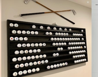 May include: A dark stained wooden display shelf filled with golf balls, mounted on a wall. Two crossed golf clubs are mounted above the shelf. The golf balls have various logos and designs.