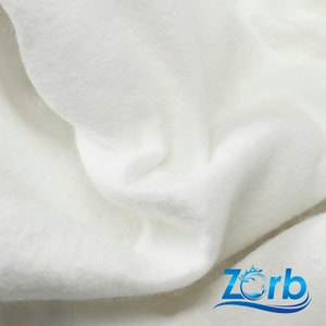 May include: Close-up of a white, soft, and fluffy fabric. The fabric is textured and has a slight sheen. The fabric is folded in a way that shows its texture and drape. The fabric is labeled with the word "Zorb" in blue text with a wave design.
