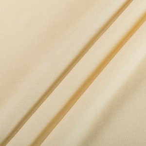 Puede incluir: Primer plano de una tela suave de color crema con una textura sutil. La tela está doblada en diagonal, creando líneas suaves y fluidas.