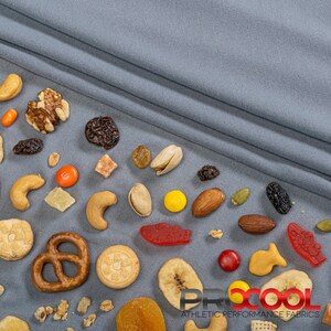 May include: A variety of snacks, including pretzels, nuts, dried fruit, and candy, are scattered on a gray fabric background. The text "PROCOOL ATHLETIC PERFORMANCE FABRICS" is visible in the lower right corner of the image.