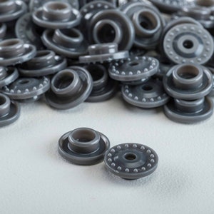 May include: A pile of gray plastic snap fasteners, with two individual fasteners in the foreground. The fasteners have a raised ring around the center hole.
