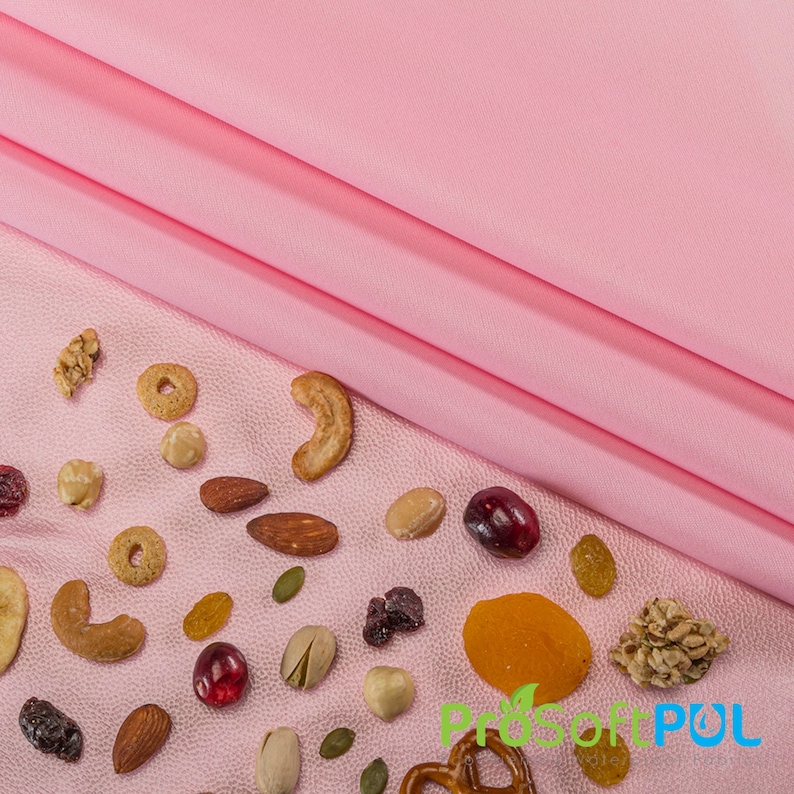 May include: A pink fabric background with a variety of nuts, dried fruit, and cereal scattered on it. The fabric is folded over in the top right corner of the image.