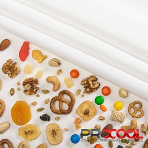 May include: A white fabric background with a variety of snacks scattered across the surface. The snacks include pretzels, nuts, dried fruit, cereal, and candy. The text "PROCOOL ATHLETIC PERFORMANCE FABRICS" is visible in the bottom right corner of the image.