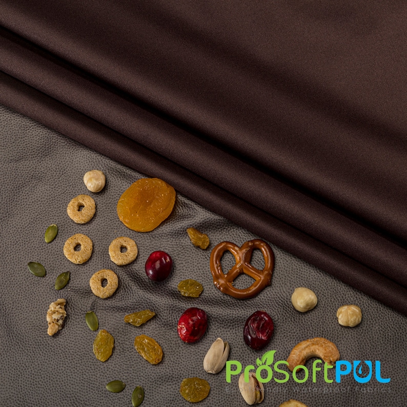 May include: A variety of snacks and dried fruit on a gray textured surface. The snacks include pretzels, dried cranberries, pistachios, cereal rings, dried apricots, and pumpkin seeds. The surface is partially covered by a brown fabric.