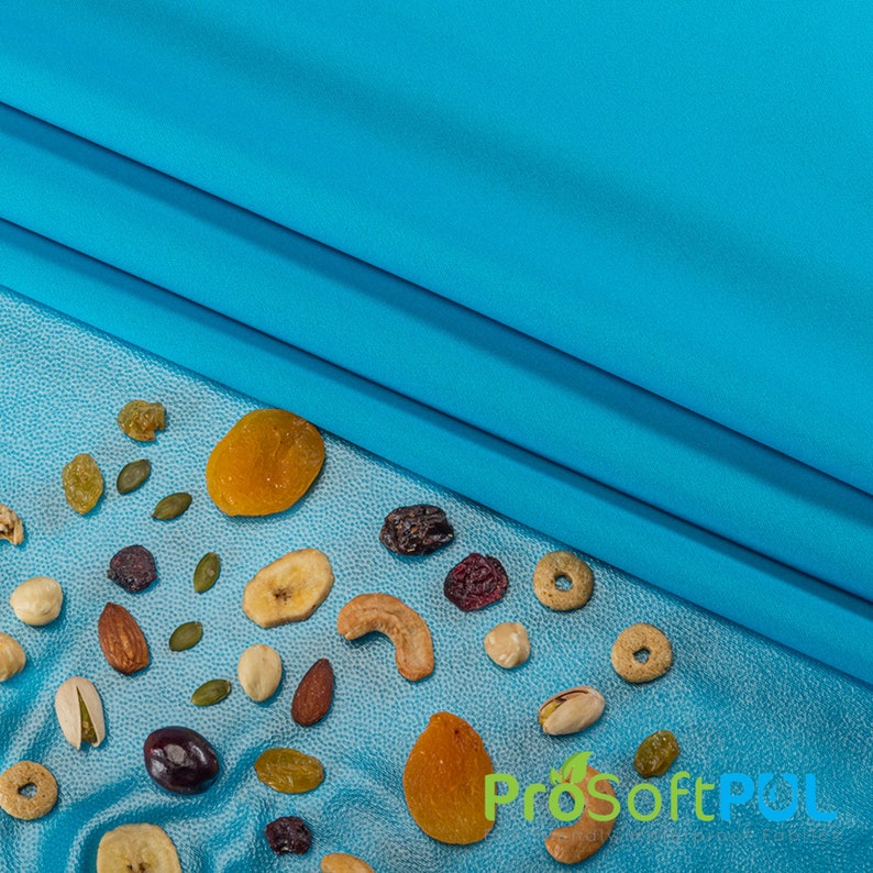 May include: A blue fabric background with a variety of dried fruits, nuts, and cereal scattered on a blue textured surface. The dried fruits include apricots, bananas, and raisins. The nuts include almonds, pistachios, and pumpkin seeds. The cereal is in the shape of small rings.