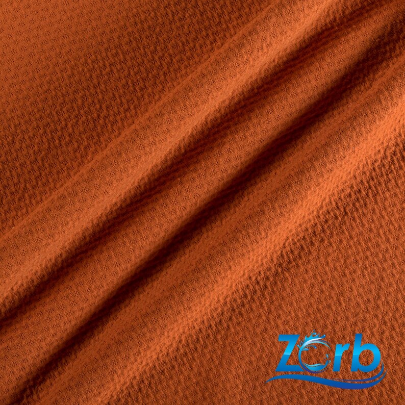 May include: Close-up of a rust-colored waffle weave fabric. The fabric has a textured surface and is folded in a diagonal pattern.