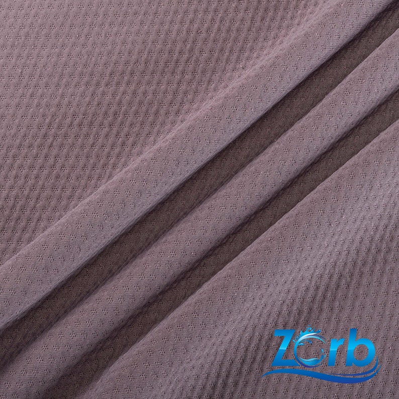 Puede incluir: Primer plano de una tela de color malva con un patr&oacute;n de tejido de gofre texturizado. La tela est&aacute; doblada en tres partes, creando una l&iacute;nea diagonal a trav&eacute;s de la imagen. La tela es suave y tiene un brillo sutil.