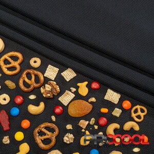 May include: A black fabric background with a variety of snacks scattered on it, including pretzels, cereal squares, nuts, candy, and dried fruit.