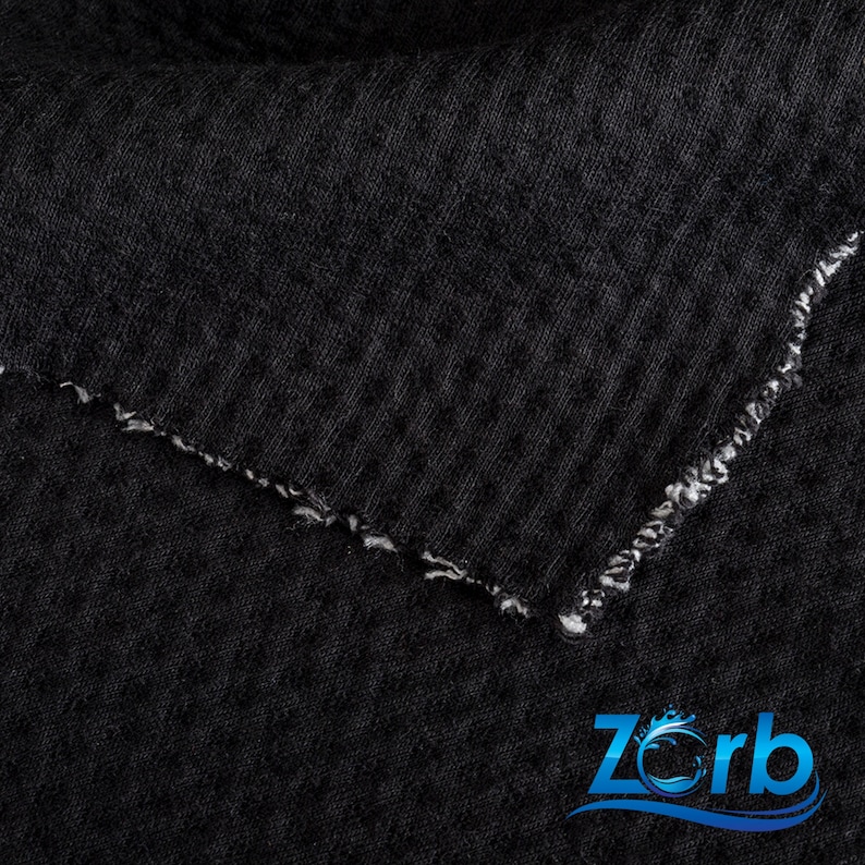 May include: Close-up of a black fabric with a textured surface. The fabric has a raw edge with white stitching.